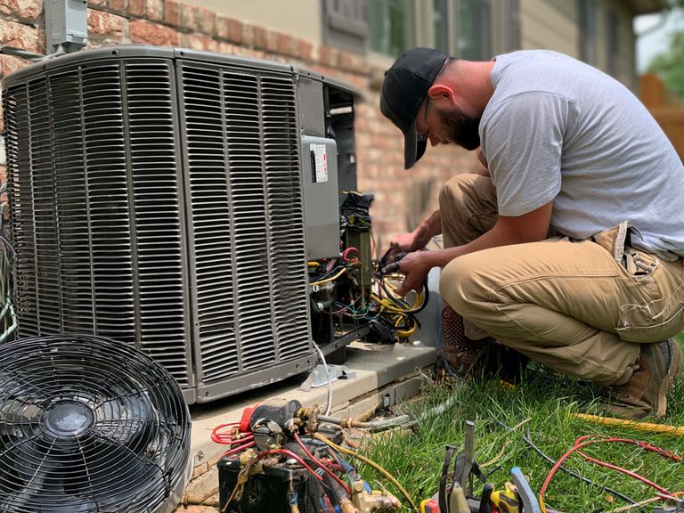 HVAC technician working on the wiring of an outdoor condenser unit, with tools laid out on the grass